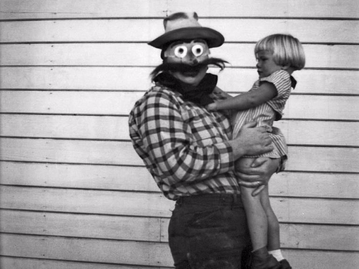 All the photos feature candid shots of family members, friends, and children in Halloween garb in the US. Here, a man with a handmade cowboy mask holds a young girl.